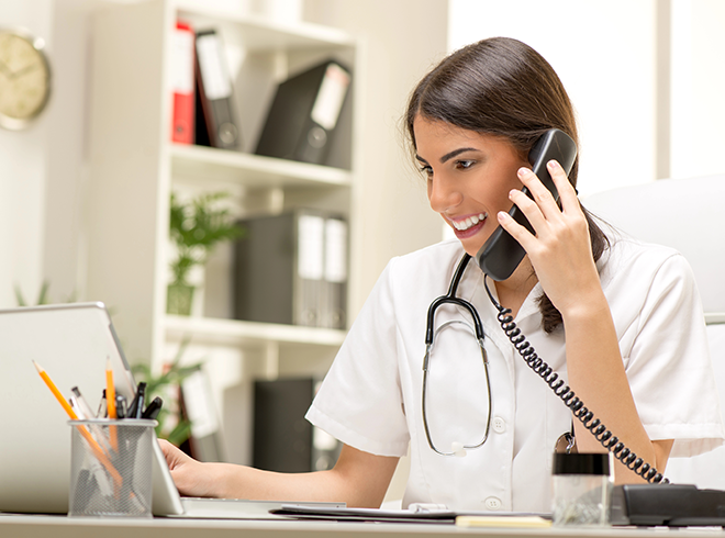 Smiling nurse on phone call while working on laptop in medical office, representing patient outreach, care coordination, and proactive clinical communication