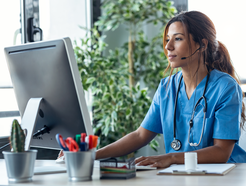 Nurse wearing scrubs and a stethoscope, speaking through a headset while working at a computer for monthly time tracking and documentation in Chronic Care Management (CCM).