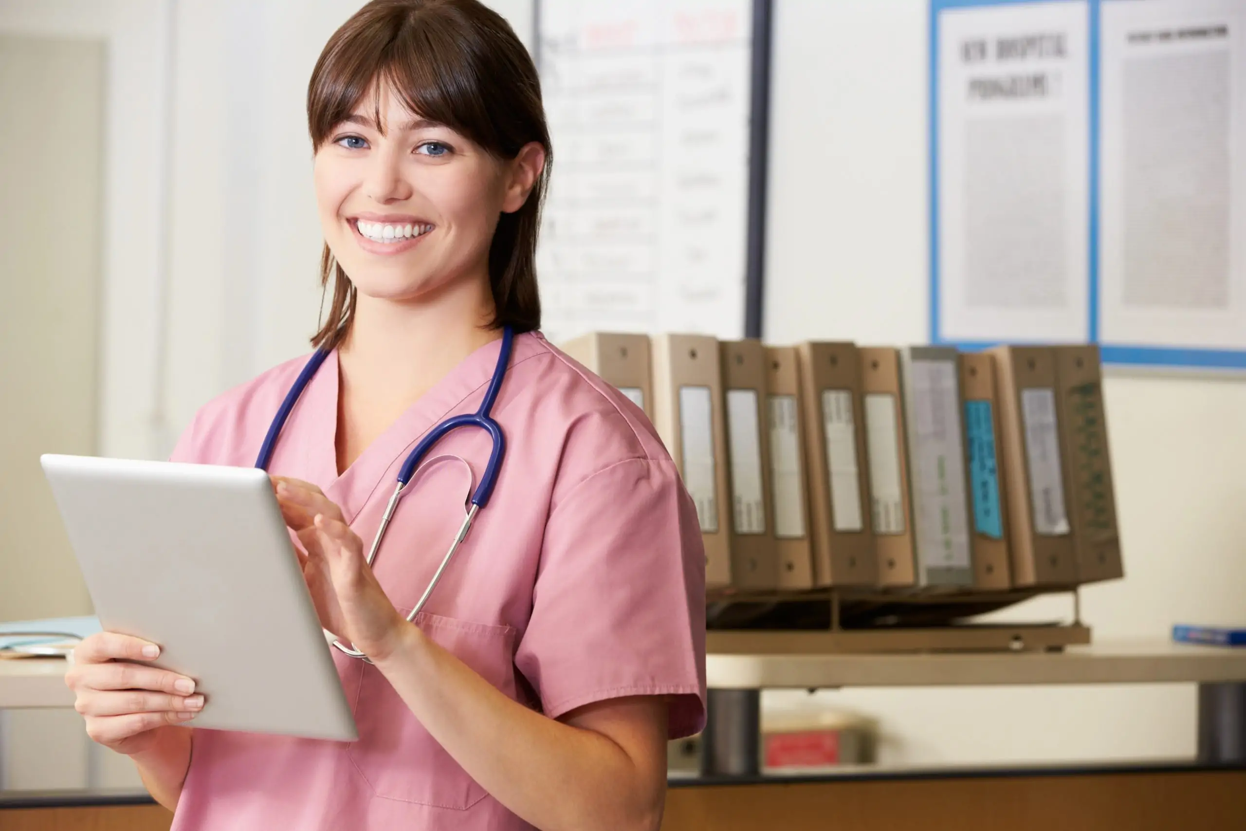 Smiling nurse in pink scrubs using a digital tablet in a medical office for patient record management.