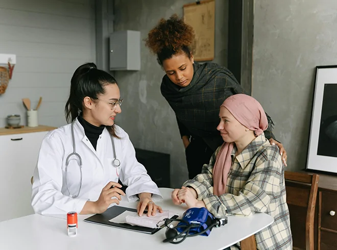 Female physician supporting cancer patient during home consultation with caregiver present, emphasizing compassionate care coordination and chronic condition management