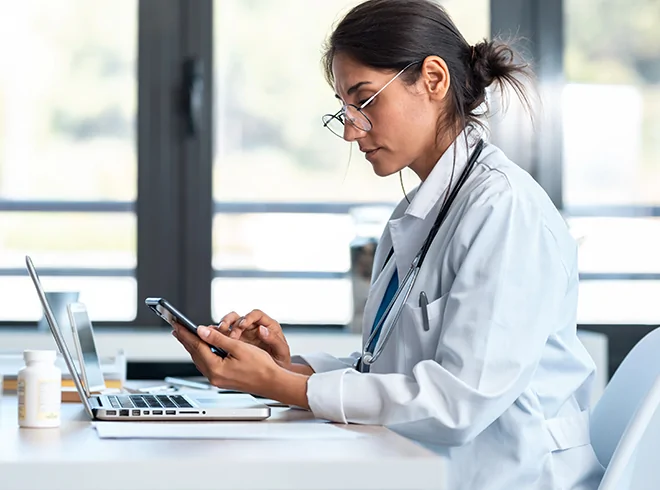 Female physician using smartphone and laptop in clinical office, illustrating digital health management, secure communication, and EHR workflow optimization