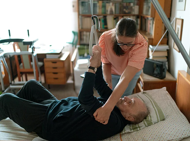 Female caregiver assisting elderly man with mobility support using a bed trapeze in a home care setting, representing functional support and aging-in-place care