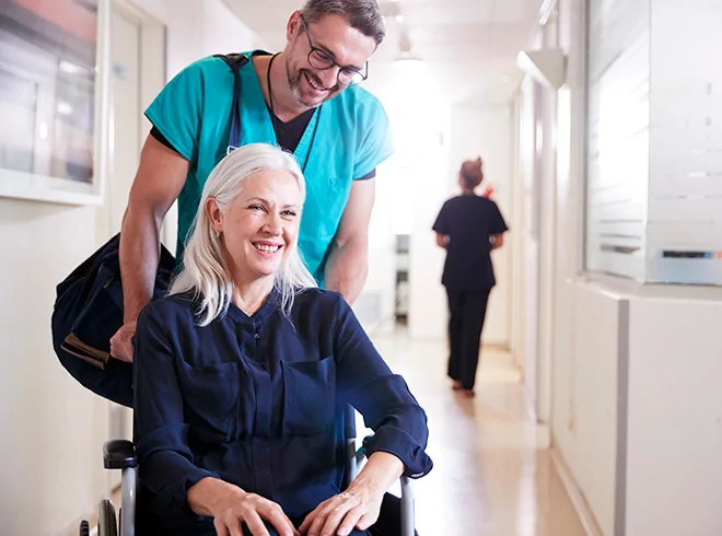 Healthcare provider assisting smiling older woman in wheelchair through clinic hallway, highlighting patient mobility support and transitional care services