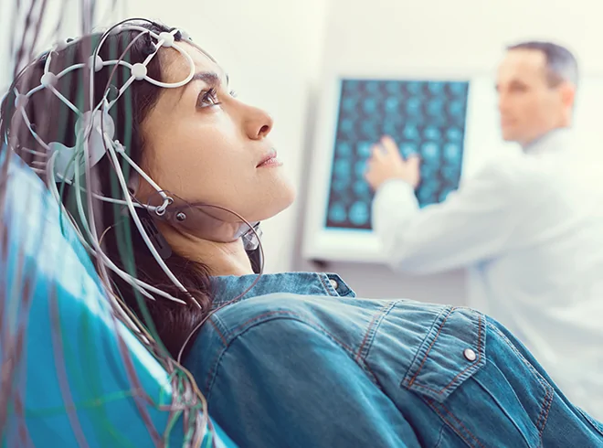 Female patient undergoing EEG test for neurological evaluation, with clinician reviewing brain scan results in background, highlighting diagnostic imaging in neuro care