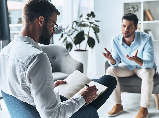 Male therapist taking notes while speaking with patient during mental health counseling session, illustrating behavioral health support and Collaborative Care Model integration