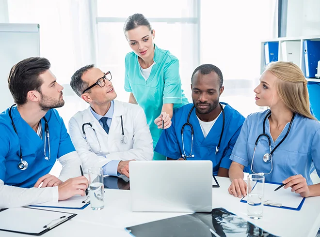 Nurse leading interdisciplinary care team discussion around laptop, illustrating nurse-led care coordination and collaborative clinical decision-making in modern healthcare settings
